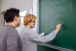 Two young men are standing in front of a blackboard on which one is writing something.