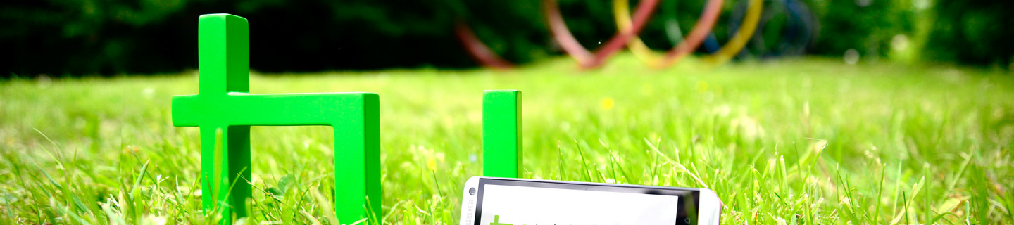The green logo of TU Dortmund University is standing on a meadow. A smartphone is leaning against it.