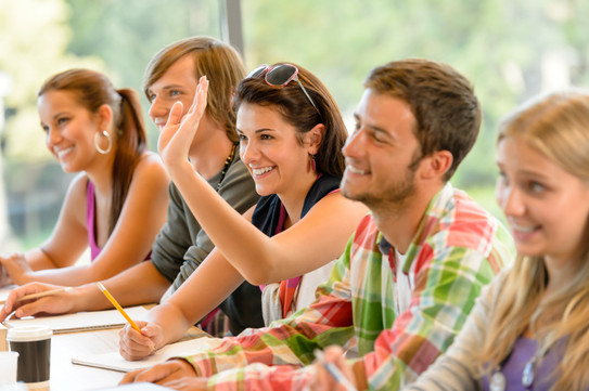 Schülergruppe Zwei Jungen und zwei Mädchen sitzen neben einander an einem Tisch im Klassenraum und heben die Hand.